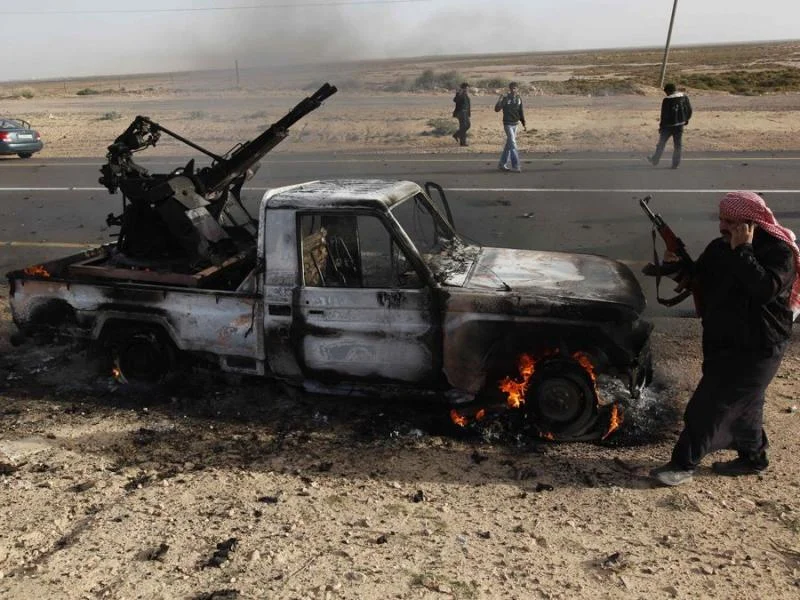 a-libyan-rebel-with-an-ak-47-walks-by-a-burned-out-pro-qaddafi-vehicle.webp